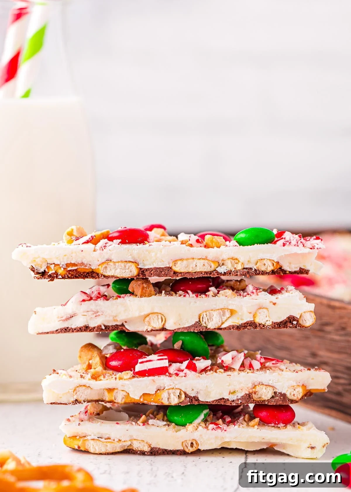 A cozy shot of four stacked pieces of Christmas bark, generously topped with pretzels, M&Ms, and crushed candy canes, with a refreshing glass of milk softly blurred in the background, suggesting a delightful treat moment.