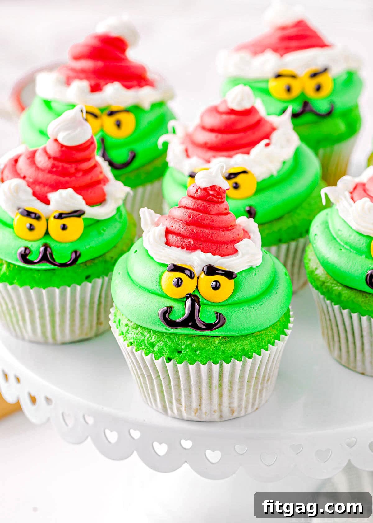 six cupcakes decorated like the grinch with red santa hats sitting on a white cake stand.