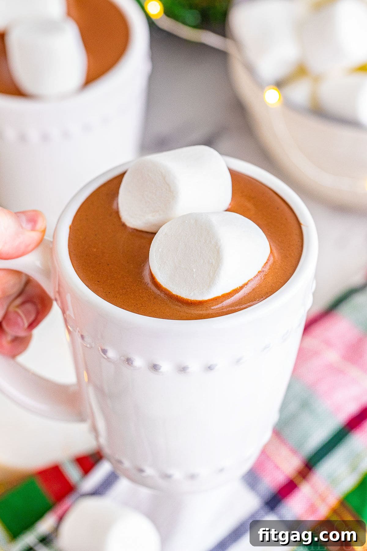 A white mug filled with luxurious crockpot hot chocolate, garnished with two jumbo marshmallows, held over a festive Christmas linen.