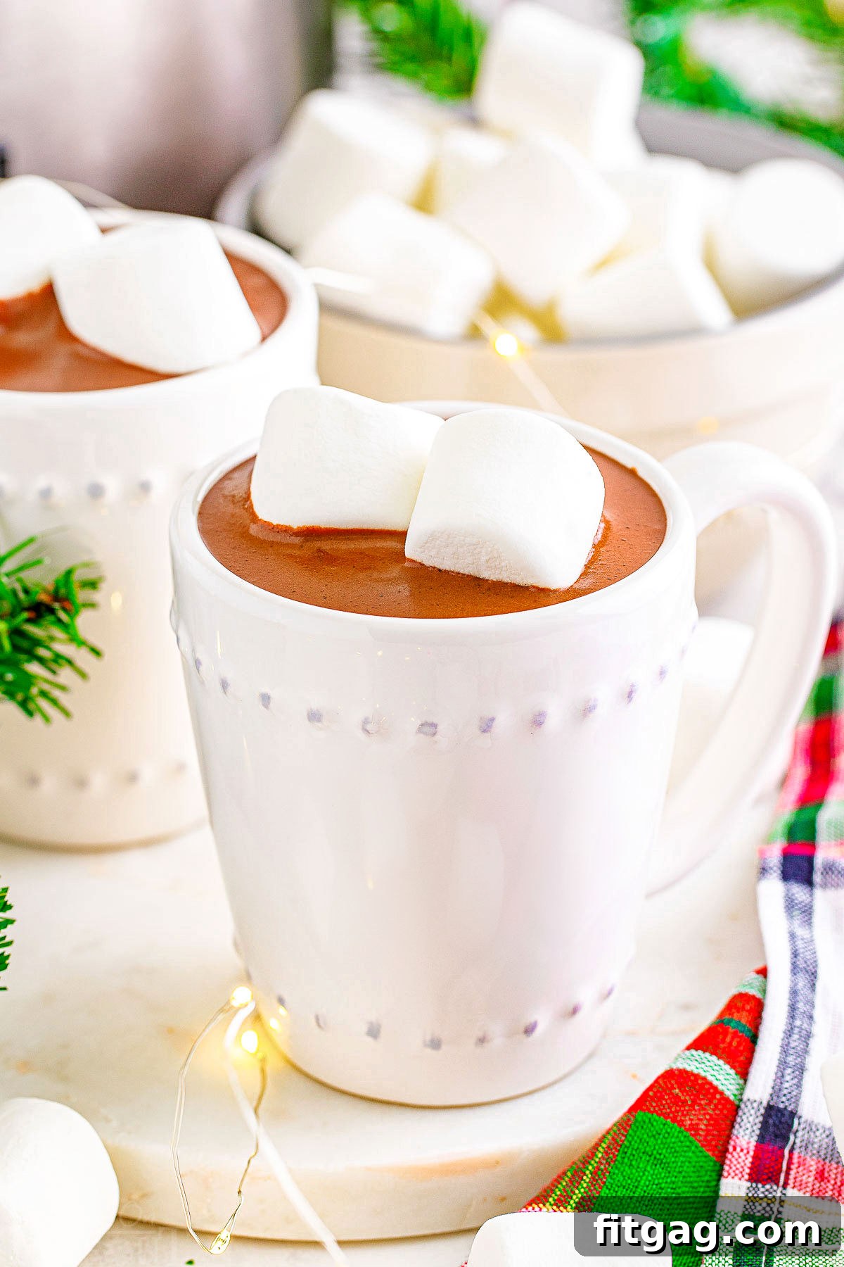 Two white mugs, one filled with hot chocolate topped with marshmallows, sitting in front of another mug of rich crockpot hot cocoa, against a rustic background.