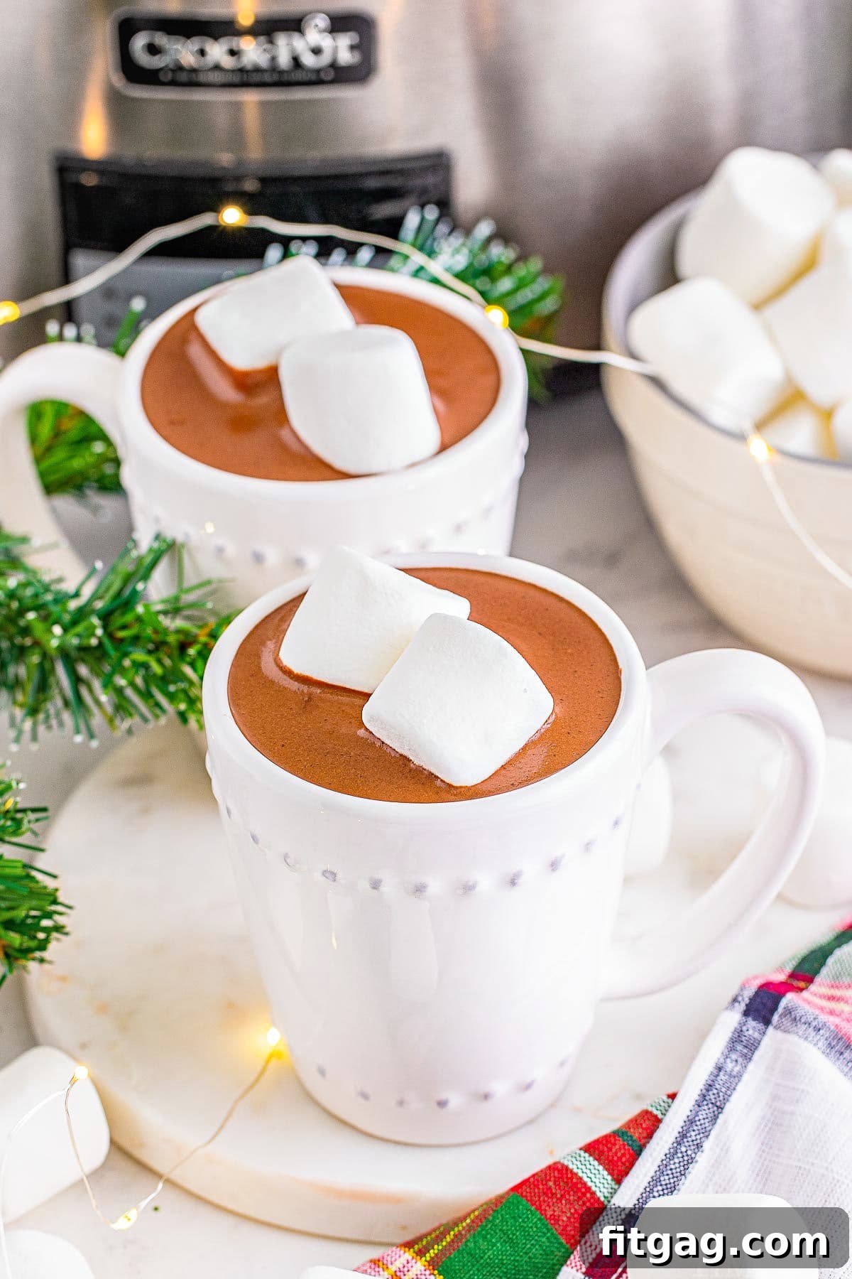Two mugs of hot chocolate, each topped with two marshmallows, are set in front of a silver crockpot, ready to be enjoyed.