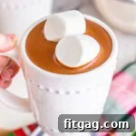 white mug filled with decadent crockpot hot chocolate and topped with two jumbo marshmallows. hand can be seeing holding the mug up and over a christmas linen.