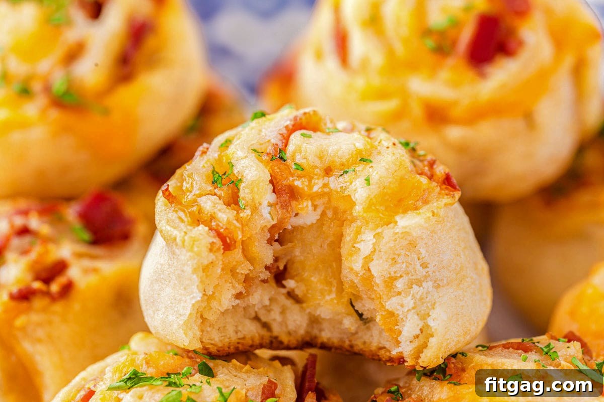 Cheesy Bacon Pinwheel Bites 3 Close-up of a Cheddar Bacon Pinwheel with a bite taken out, resting on a stack of freshly baked pinwheels, highlighting its flaky texture and cheesy filling.
