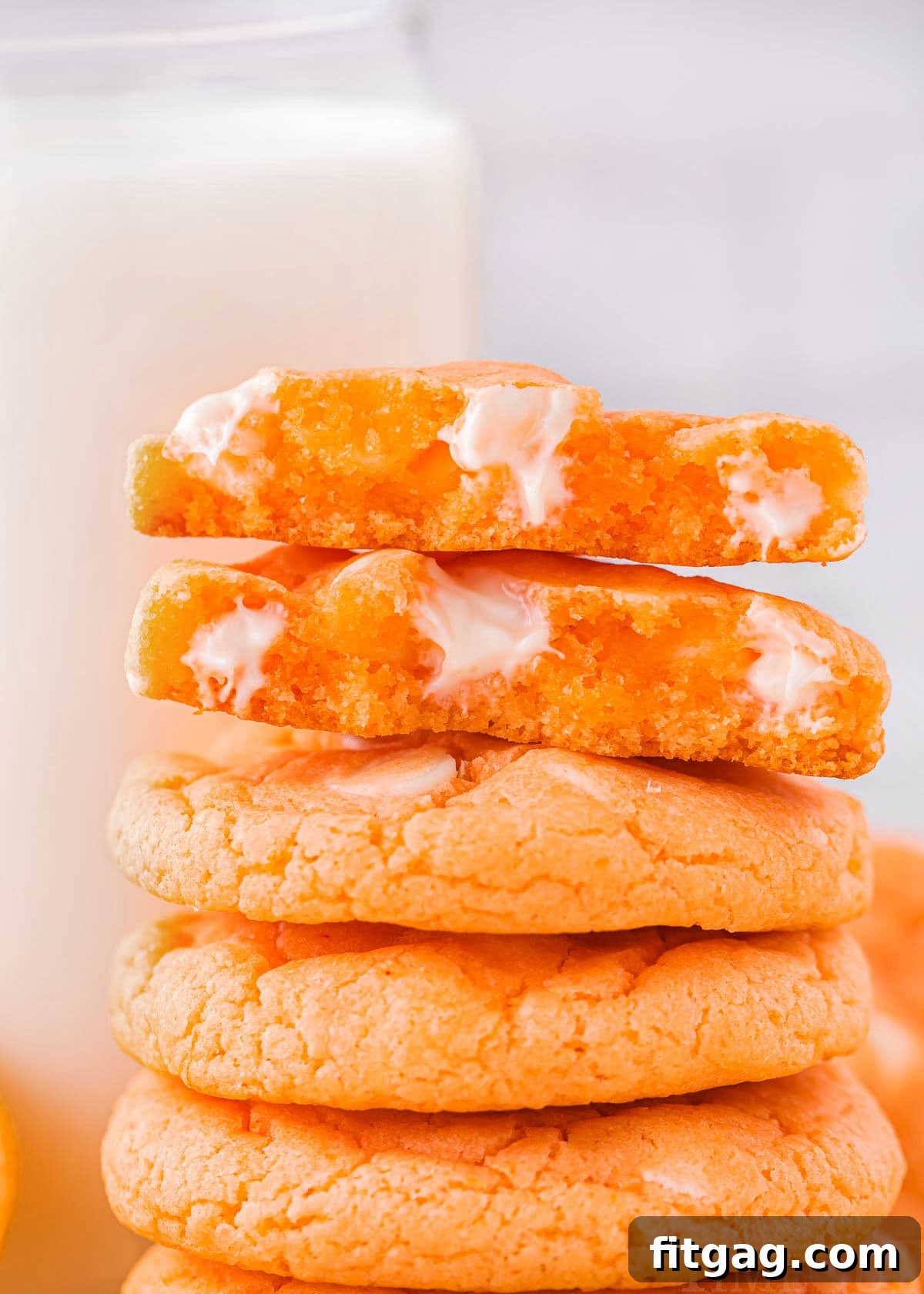 Five soft and chewy orange creamsicle cookies stacked neatly with a glass of milk in the background. The top cookie is broken in half, revealing gooey white chocolate chips melted within the cookie's tender interior.
