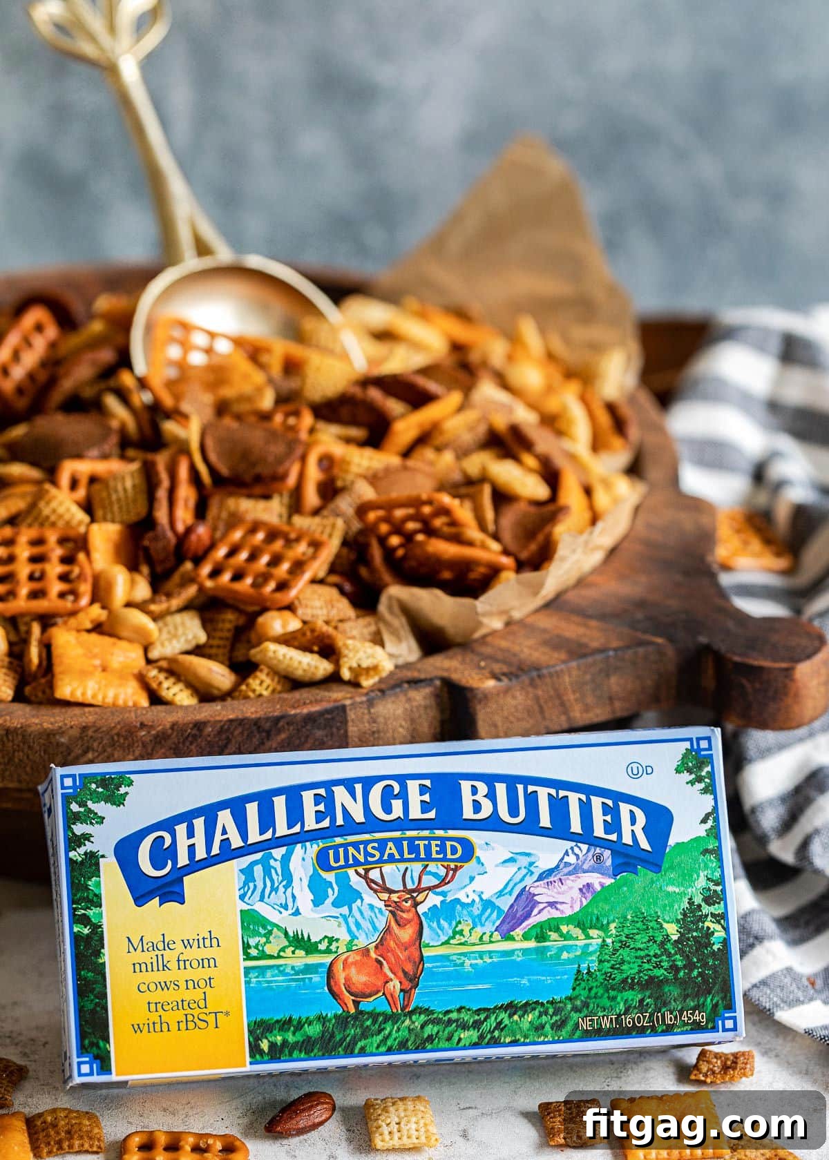 A box of Challenge butter positioned in front of a large wooden bowl filled with Chex Mix, with a gold scoop peeking out.