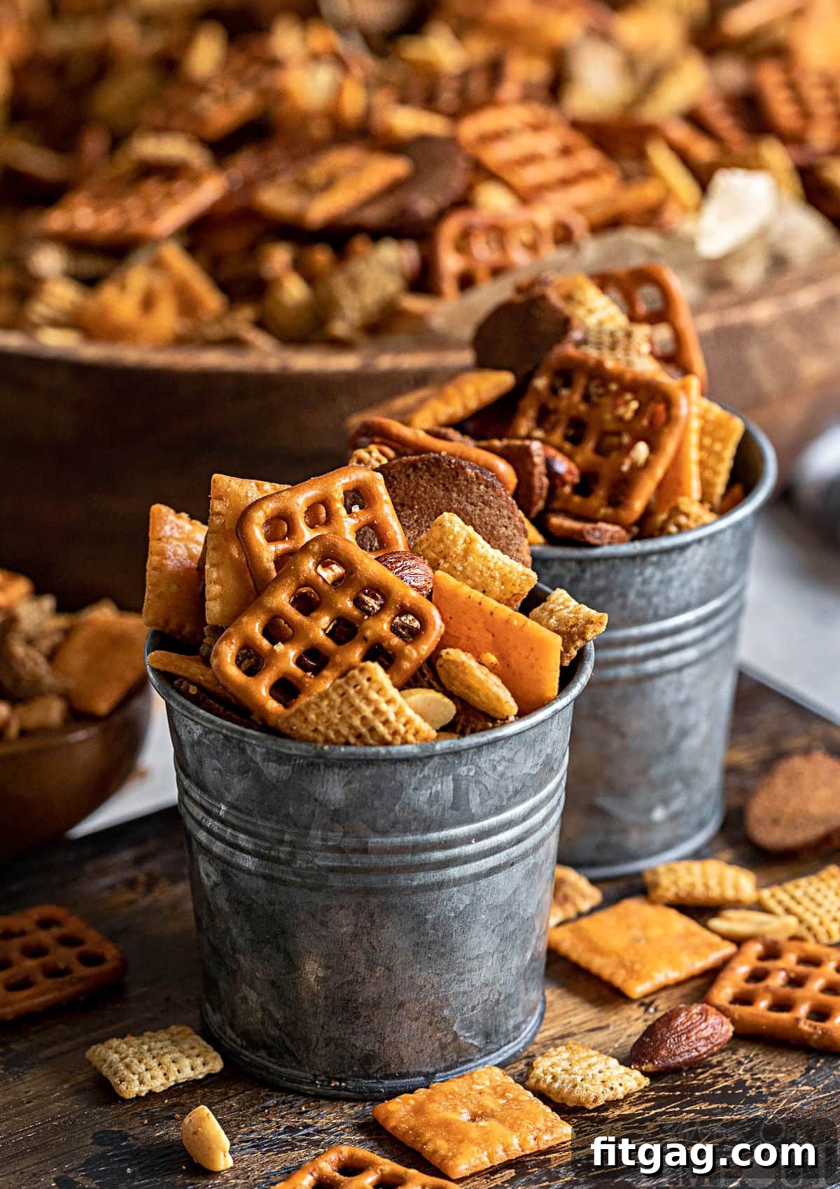 Chex Mix served in small metal containers with a large wooden bowl of the remaining mix in the background, showcasing its delicious appeal.