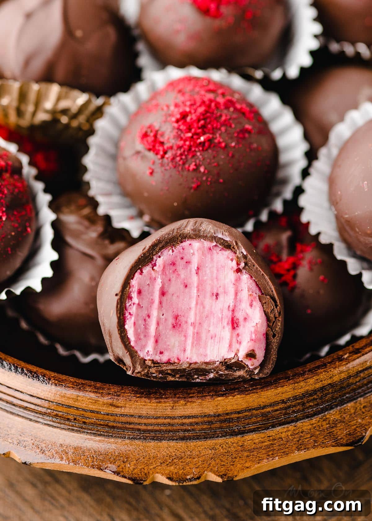A charming round wooden box brimming with homemade raspberry truffles. One truffle in the foreground has a bite taken out of it, revealing its creamy, vibrant pink interior. Several truffles are delicately dusted with freeze-dried raspberry powder for an elegant finish.