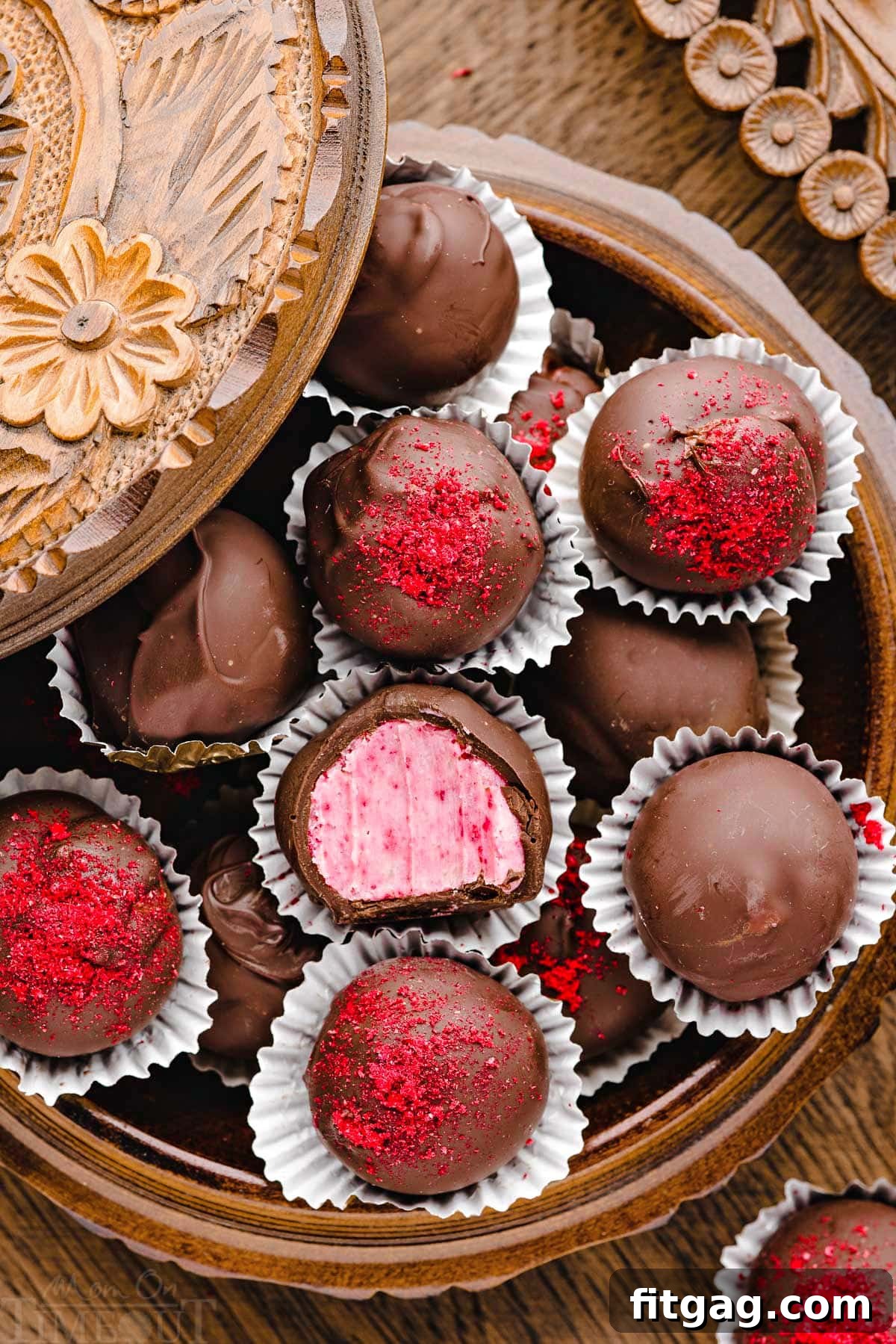 A beautifully arranged round wooden box filled with an assortment of raspberry truffles. The truffle in the center has a clean bite removed, clearly showing the distinct layers of rich chocolate coating and a creamy, pastel pink raspberry ganache interior. Some truffles are garnished with raspberry dust.