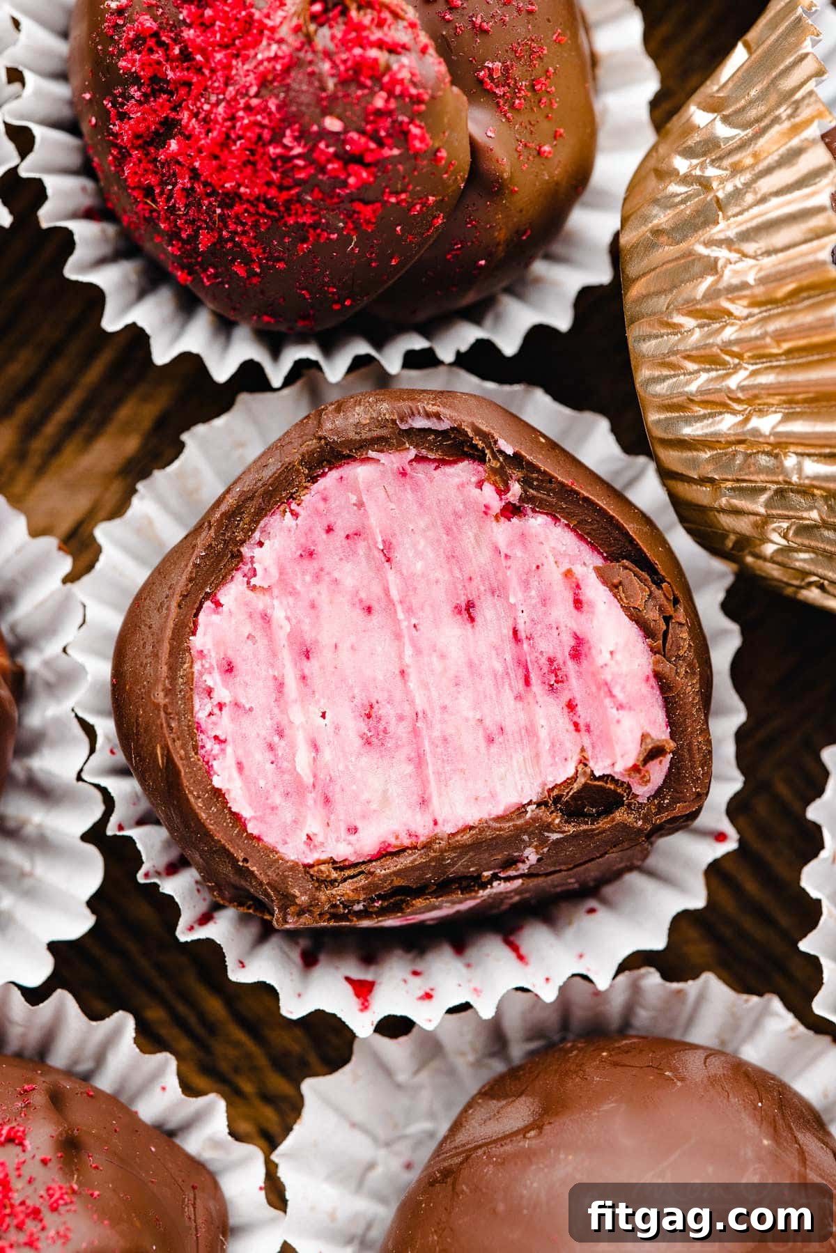 A close-up photograph of a single raspberry truffle, perfectly coated in dark chocolate and nestled in a small paper liner. Several more truffles are visible in the soft-focus background, suggesting a delightful array.
