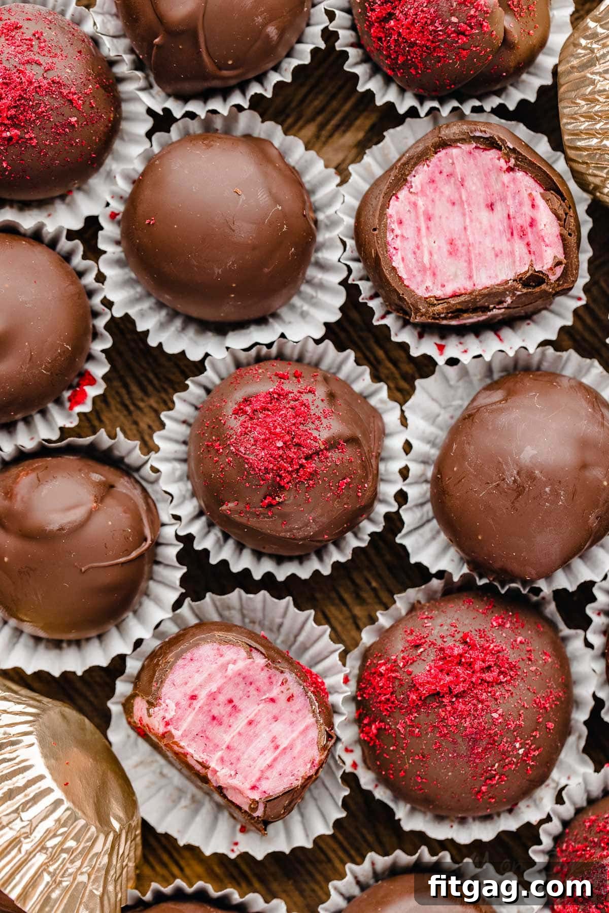 A top-down view of a dozen or so exquisite raspberry chocolate-covered truffles nestled within a rustic wooden box. Two of the truffles are artfully bitten in half, revealing their vibrant, creamy pink interiors.