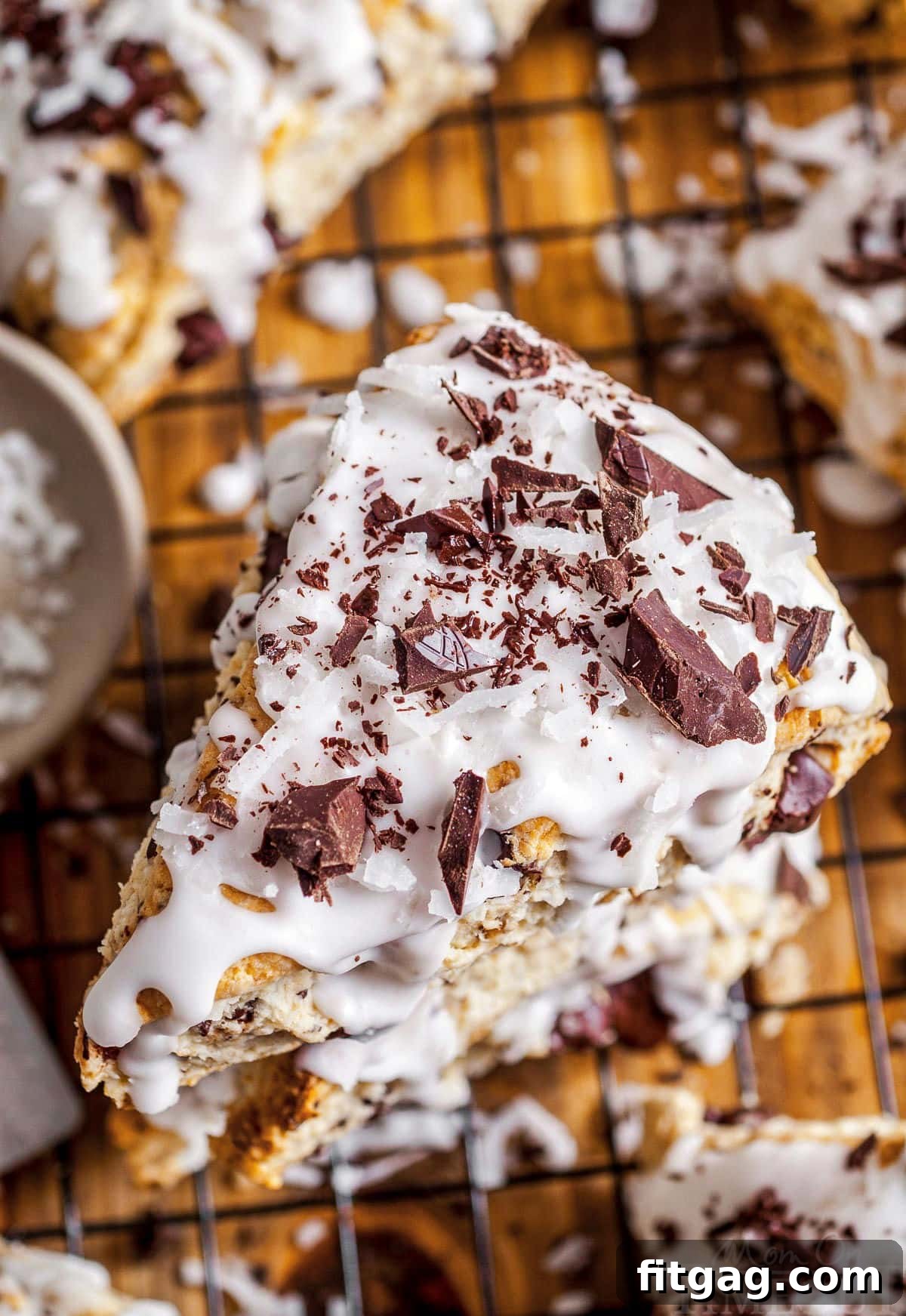 Three golden brown chocolate coconut scones, generously topped with creamy white coconut icing and dark chocolate shavings, stacked invitingly on a simple white plate. The image captures the tender texture and delicious topping, emphasizing the rich flavors of coconut and dark chocolate.