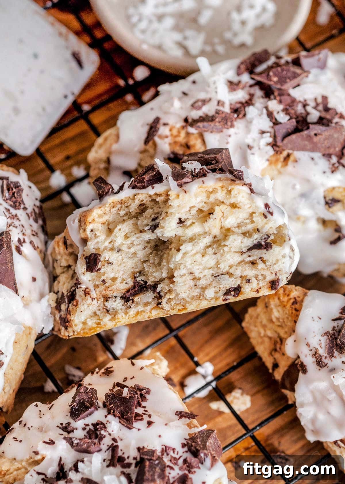 A freshly baked chocolate coconut scone, broken in half to reveal its tender, moist interior dotted with dark chocolate chunks. It sits on a wire cooling rack next to other whole scones, showcasing the delicious texture and rich ingredients.