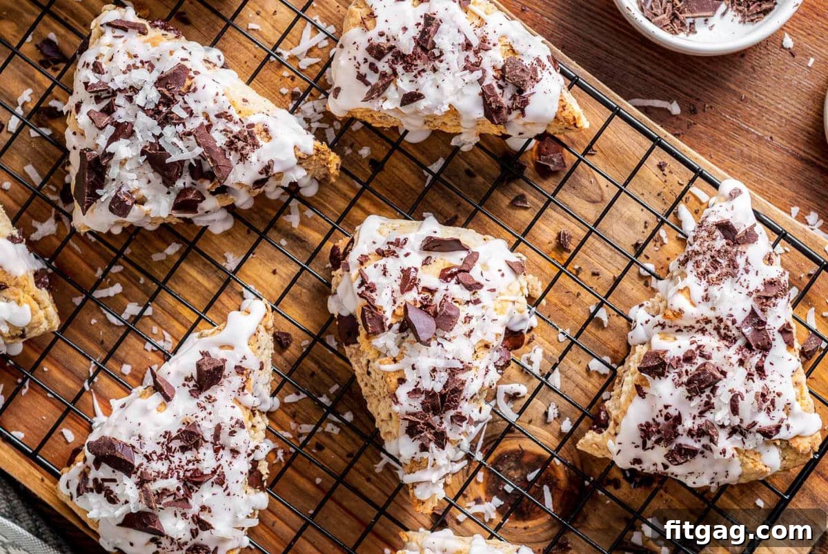 A top-down view of five beautifully iced chocolate coconut scones, garnished with chocolate shavings and coconut flakes, resting on a cooling rack atop a rustic wooden board. The scene highlights the delicious final presentation of the Almond Joy-inspired treats.