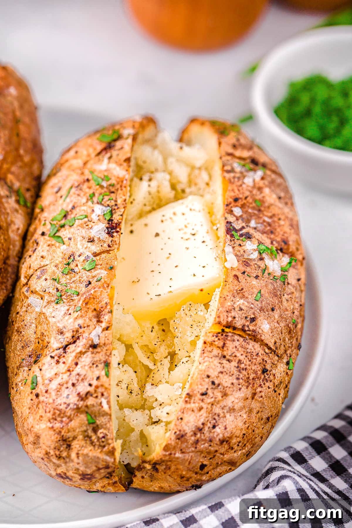 Air fryer baked potato on a white plate topped with butter, salt, pepper, and fresh green chives. A small white bowl of chopped chives is in the background.