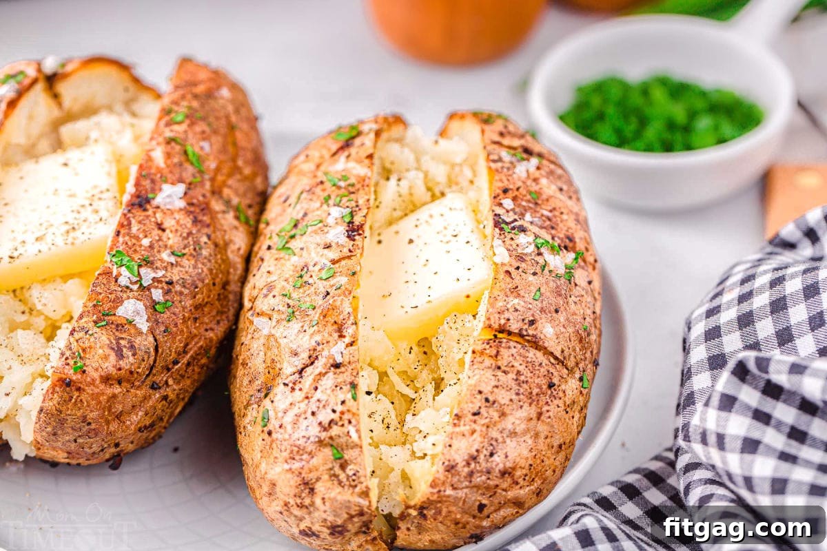 Two air fryer baked potatoes on a white plate, garnished with melting butter, a sprinkle of salt and pepper, and fresh green chives. A small bowl of chives is in the background.