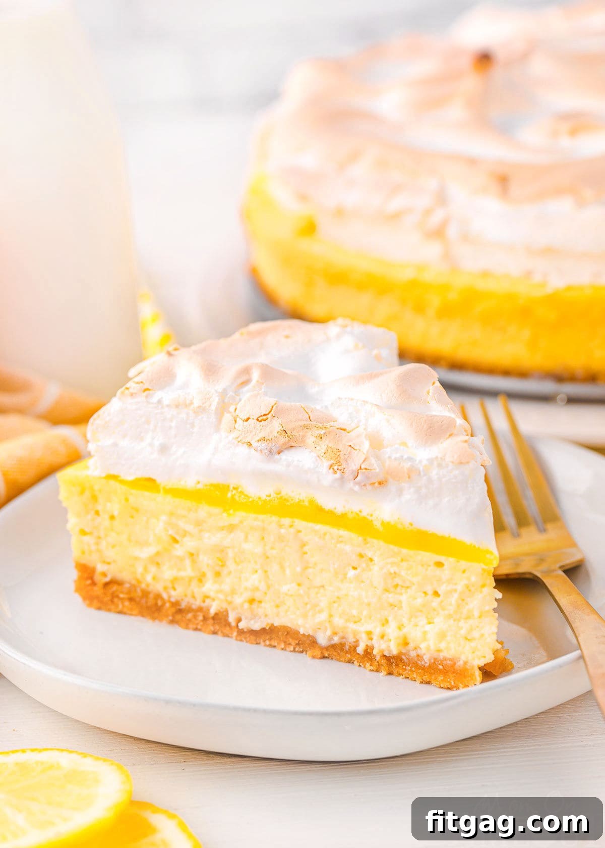 A perfectly sliced piece of lemon meringue cheesecake on a white plate, with the rest of the cheesecake and lemon slices in the background.