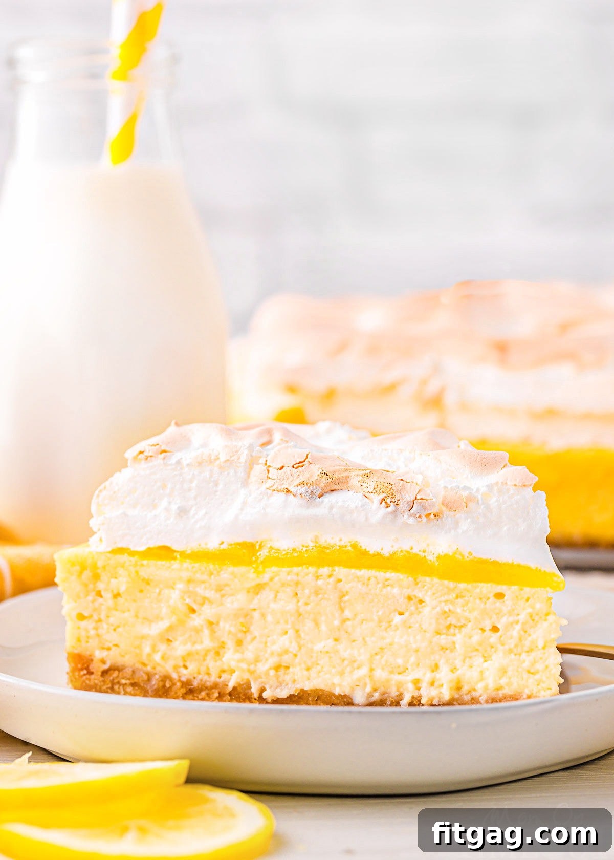 A single slice of lemon meringue cheesecake on a white plate, with the rest of the cheesecake visible in the background, accompanied by lemon slices.