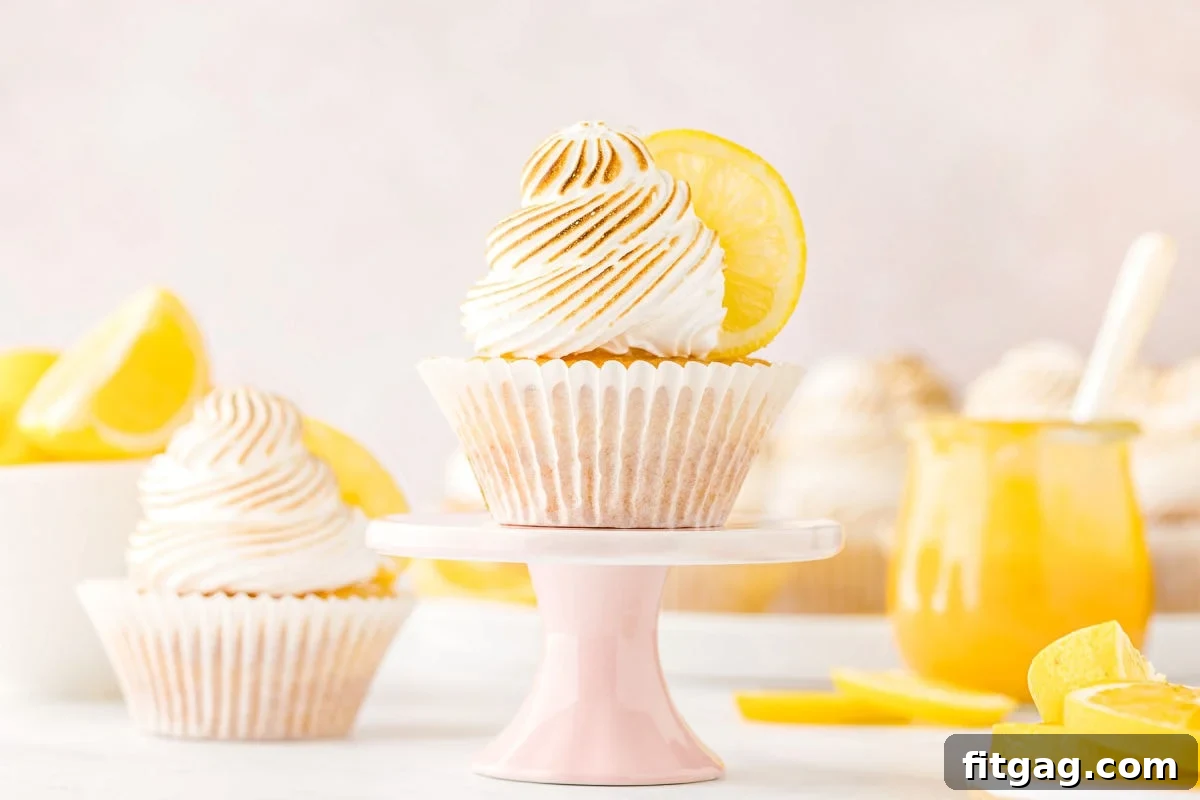Tangy Meringue Cloud Cupcakes 3 Two beautifully piped lemon meringue cupcakes, one resting on a charming pastel pink stand, with a jar of homemade lemon curd visible in the softly blurred background.