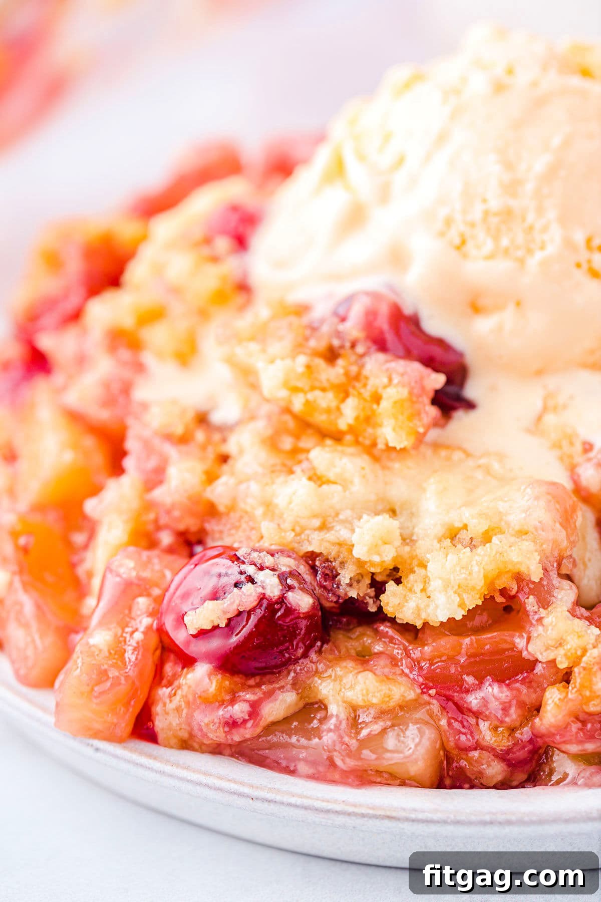 A plate filled with a slice of Cherry Pineapple Dump Cake, adorned with a melting scoop of vanilla ice cream. Visible cherries and pineapple chunks are nestled within the moist cake.