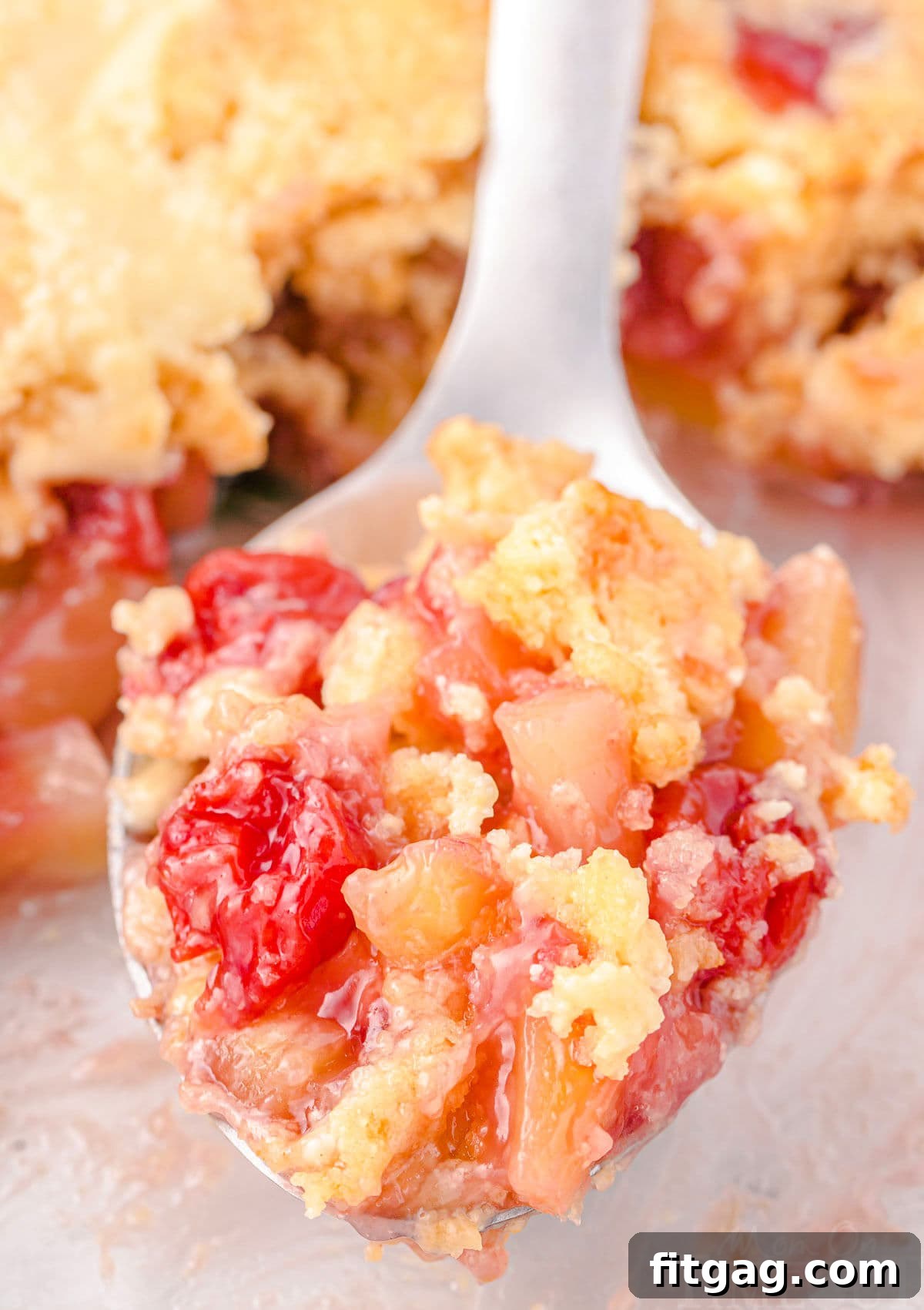 A serving spoon filled with a scoop of cherry pineapple dump cake, featuring vibrant cherry and pineapple pieces, held just above the baking dish.