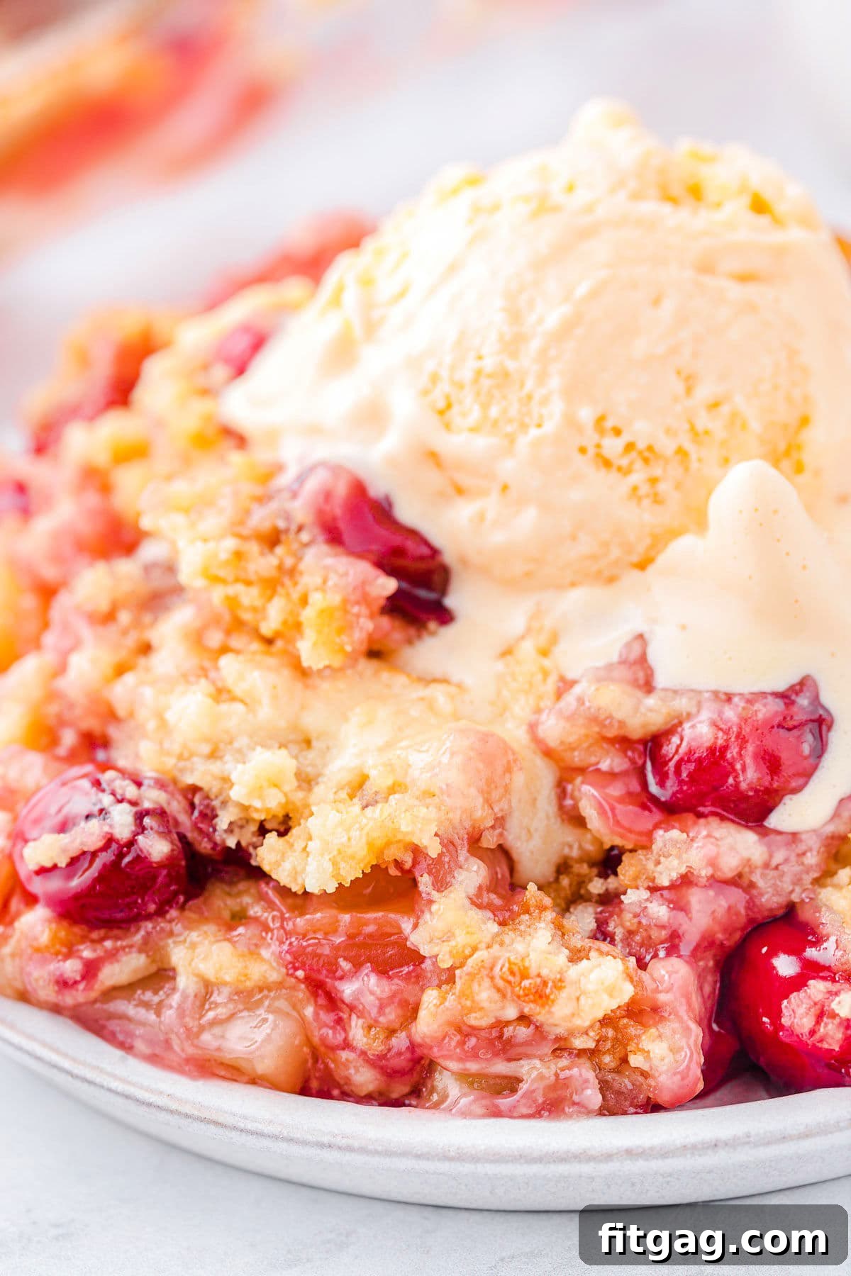 A plate filled with a slice of Cherry Pineapple Dump Cake and topped with a scoop of vanilla ice cream, which has just begun to melt, revealing delicious cherries and pineapple chunks in the cake.