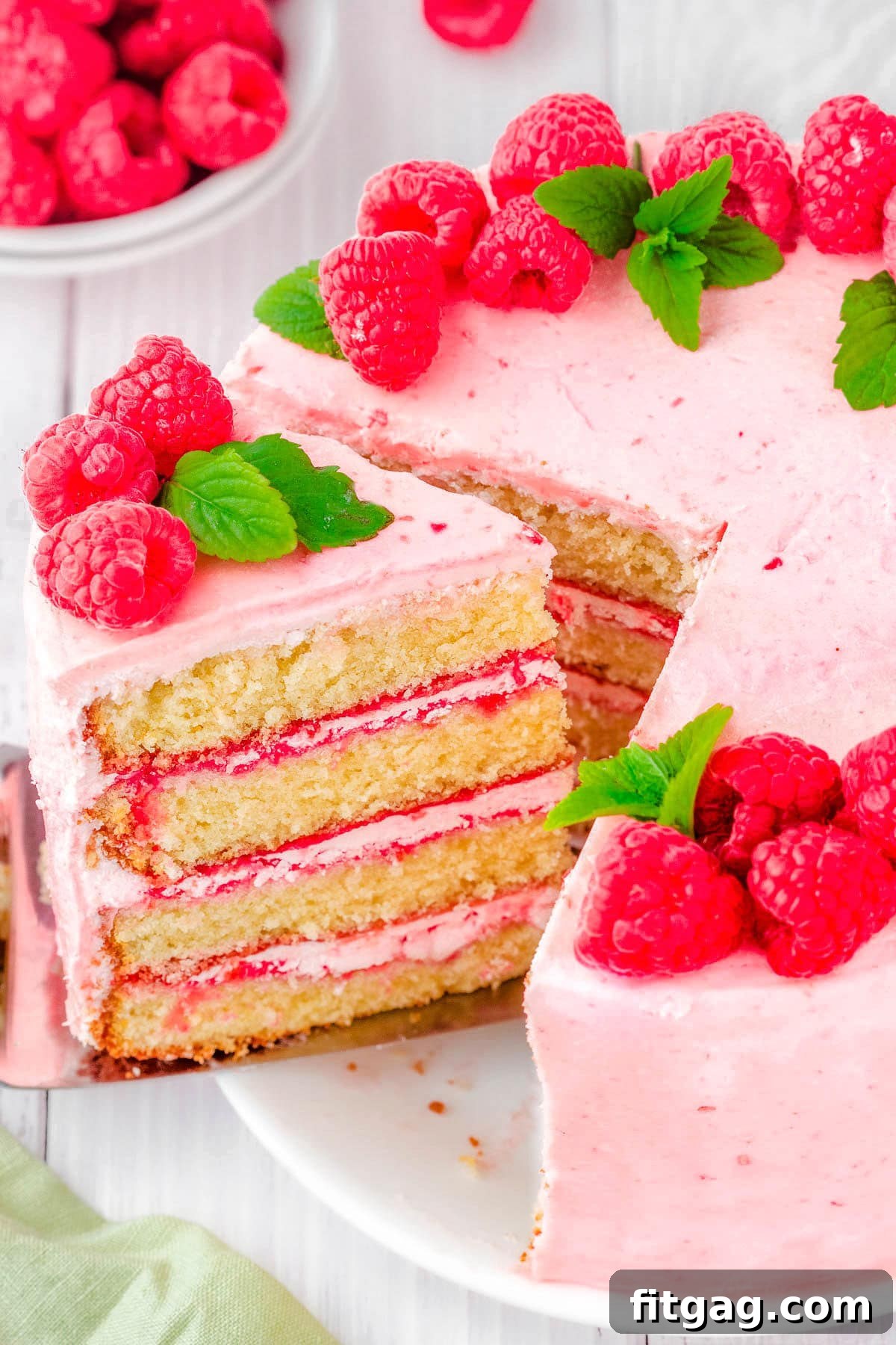 Luscious Raspberry Vanilla Cake 2 A beautiful slice being removed from a tall, four-layer vanilla cake adorned with raspberry buttercream. The cake is elegantly decorated with fresh raspberries and delicate mint leaves, showcasing its inviting layers and perfect presentation.