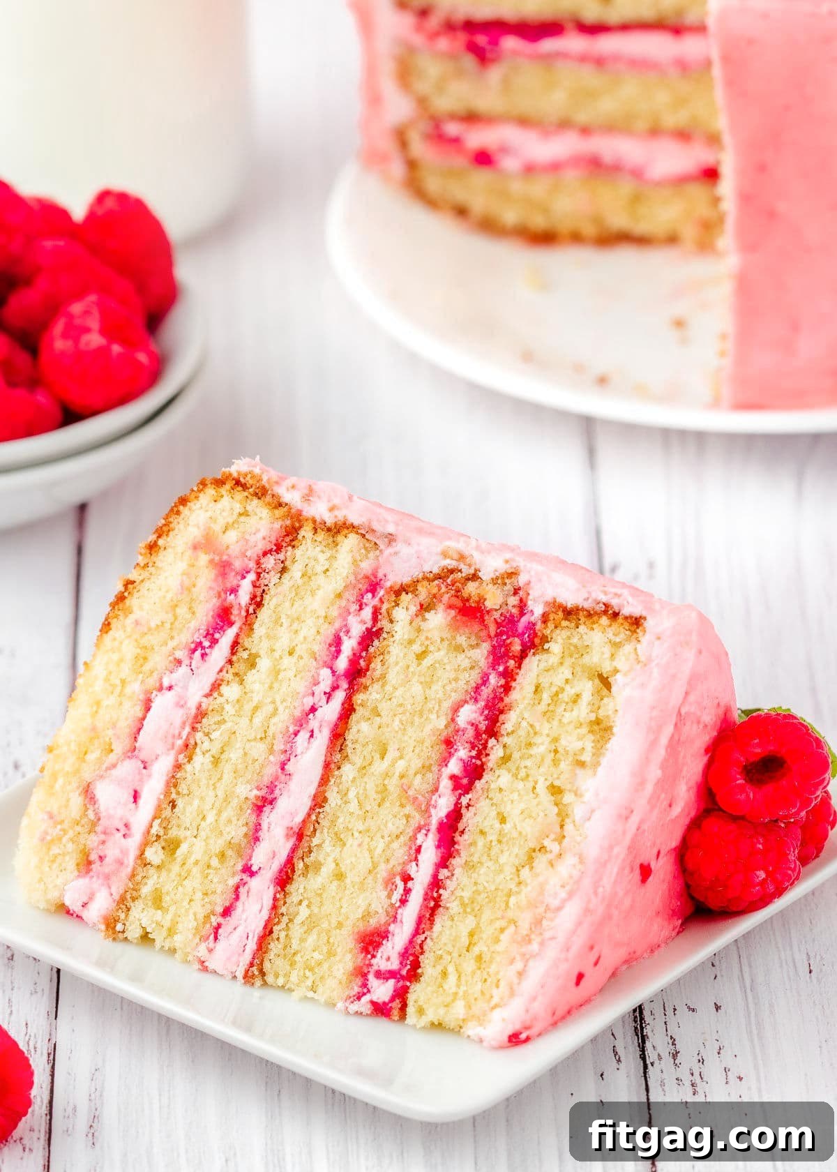 Luscious Raspberry Vanilla Cake 4 A pristine slice of vanilla raspberry cake, elegantly plated on a white square dish, positioned in front of the remaining larger cake. The image highlights the delicate layers and vibrant filling.