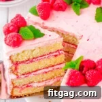 A delectable slice being removed from a vanilla cake adorned with rich raspberry buttercream. The cake is beautifully decorated with fresh raspberries and delicate mint leaves, highlighting its inviting layers.