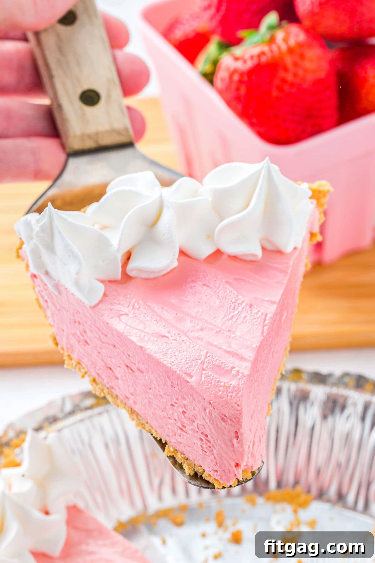 A perfectly sliced piece of vibrant Kool Aid pie, generously topped with fluffy whipped cream, is held aloft, showcasing its creamy texture and inviting color. The rest of the pie rests in the background, ready to be enjoyed.