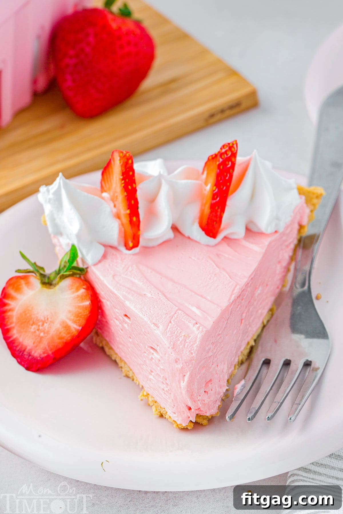 A vibrant slice of strawberry Kool Aid pie, beautifully topped with a swirl of whipped cream and fresh strawberry slices, rests on a small, elegant white plate beside a fork, ready to be savored.