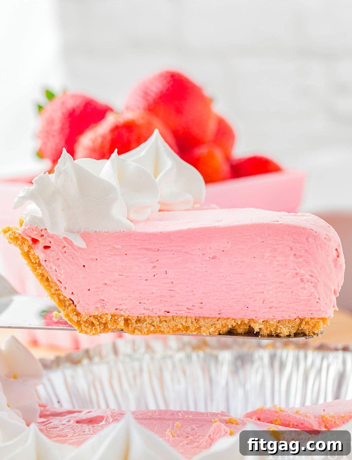 A vibrant pink slice of Kool Aid pie, perfectly topped with fresh strawberries and a swirl of whipped cream, is being carefully held by a server over the rest of the pie, ready to be presented.