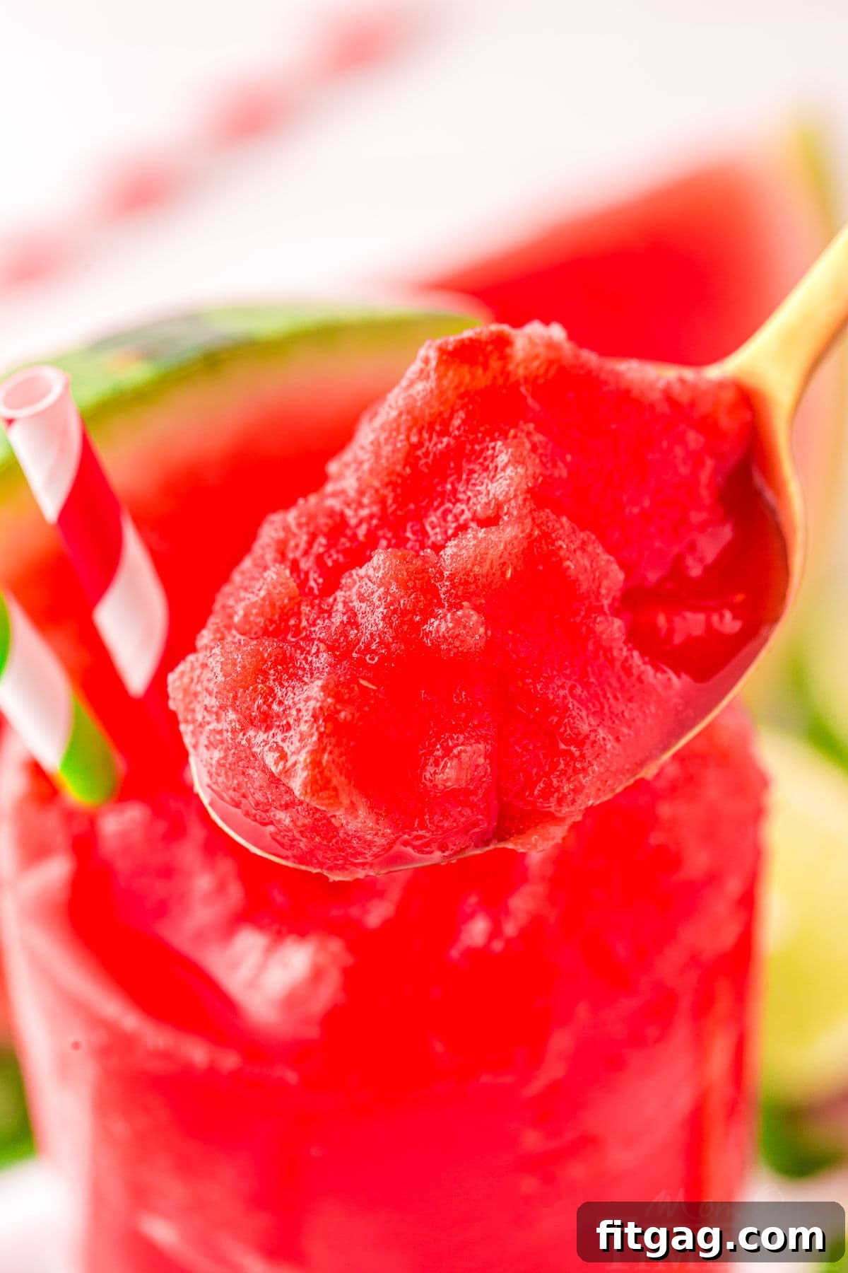 A spoon lifting a scoop of bright red watermelon slushie from a tall glass, highlighting its frosty texture.