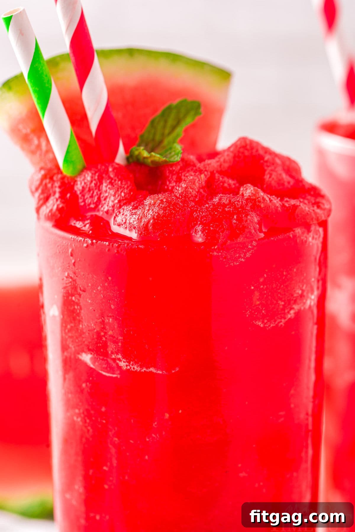 Close-up of a tall clear glass filled to the brim with vibrant red watermelon slushie, garnished with a fresh mint sprig and a watermelon wedge, with two paper straws.