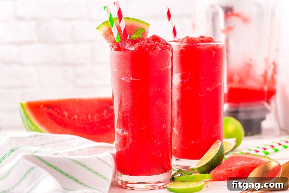 Two tall glasses of bright red watermelon slushie, garnished with watermelon wedges, with a blender and more watermelon in the background.