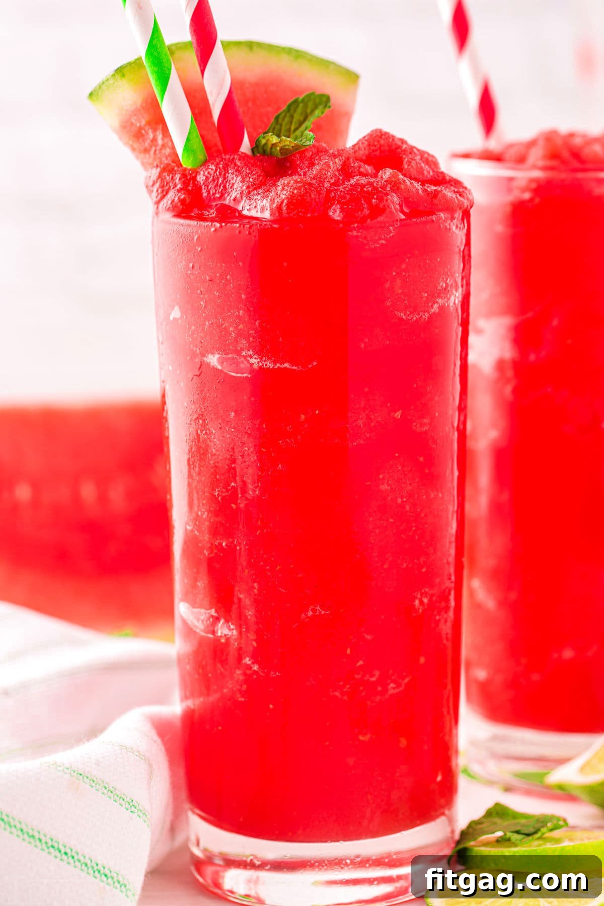 Two tall glasses of bright red watermelon slushie, garnished with watermelon wedges, glistening under warm light.