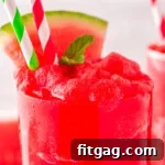 Close up view of a tall clear glass filled to the top with watermelon slushie. Topped with a sprig of mint and watermelon wedge with two paper straws inserted.