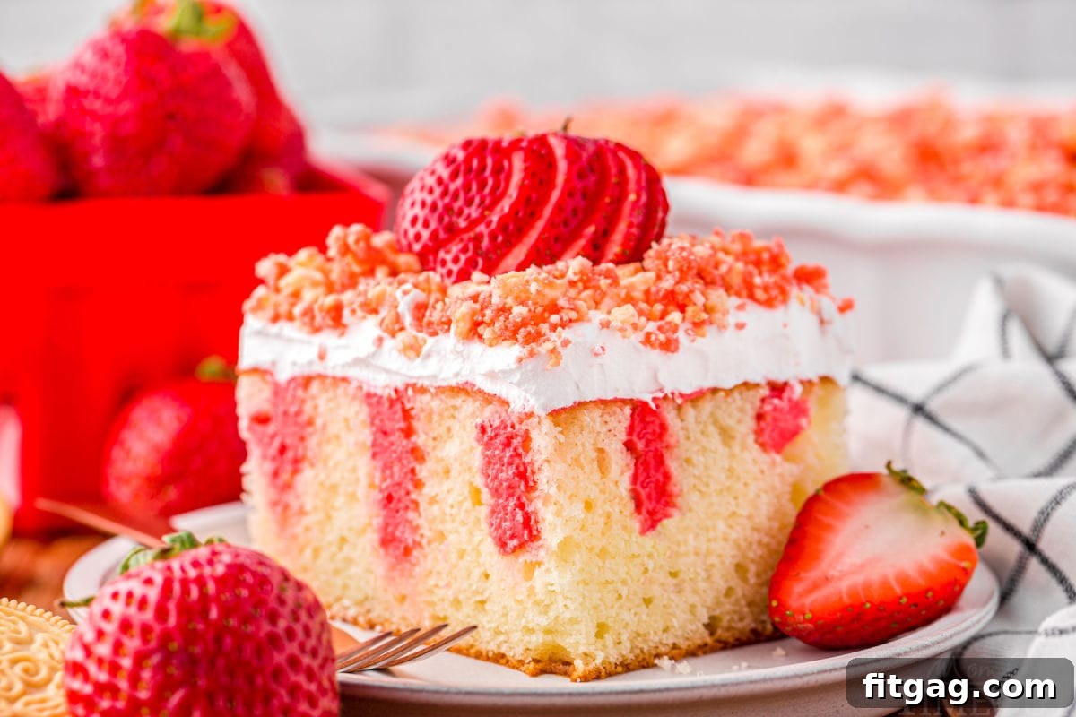 serving of strawberry poke cake on small white round plate topped with fresh sliced strawberry and crunch topping. rest of cake can be seen in background.
