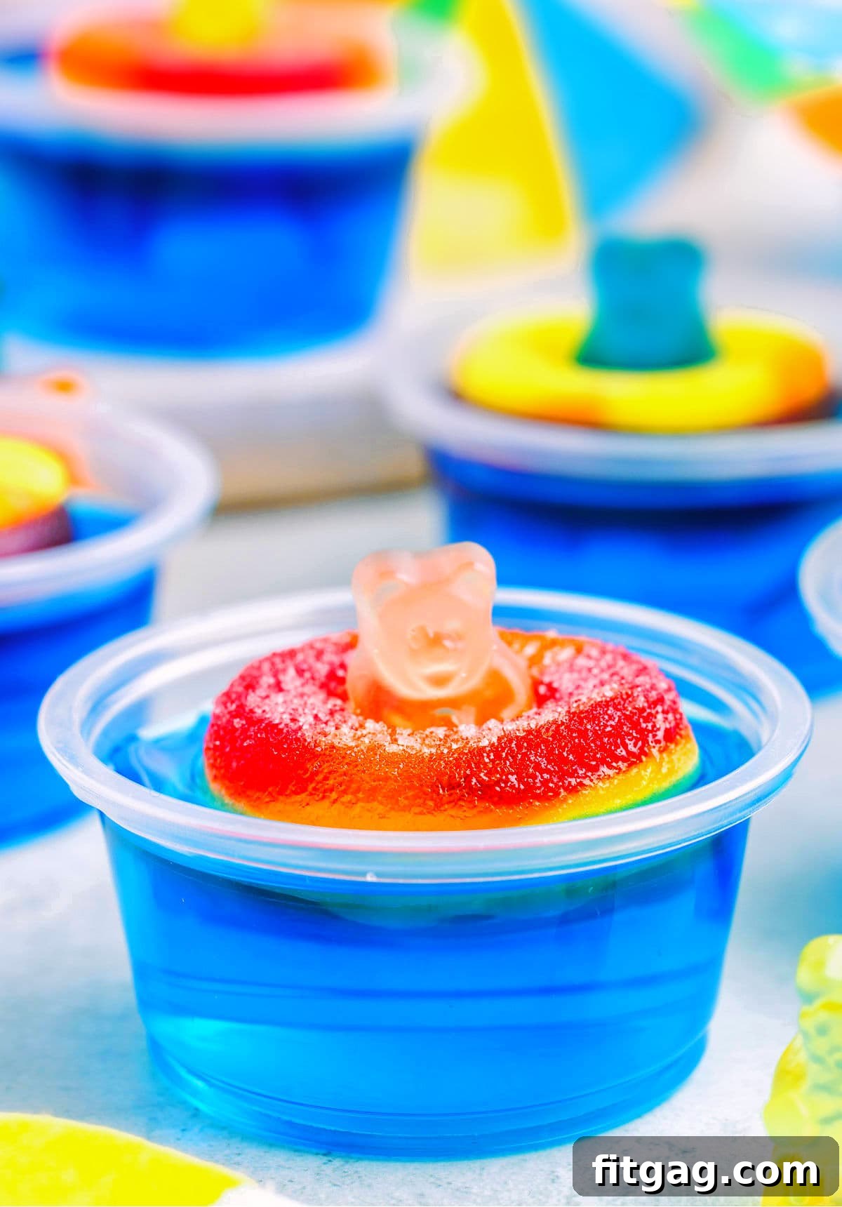 Poolside Jell-O Jiggle 2 A vibrant blue pool party jello shot featuring a happy gummy bear relaxing on a peach ring floatie, set against a pristine white background. More jello shots are visible in the soft focus background, creating a festive atmosphere.