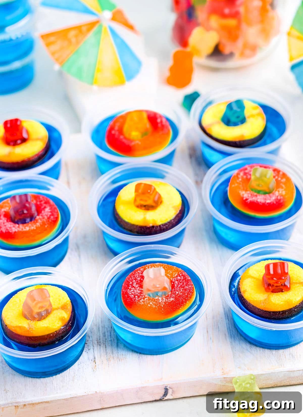 Poolside Jell-O Jiggle 3 A charming white tray adorned with multiple blue pool party jello shots, each topped with a gummy bear on a peach ring floatie. Scattered paper umbrellas and additional gummy bears enhance the whimsical party ambiance.