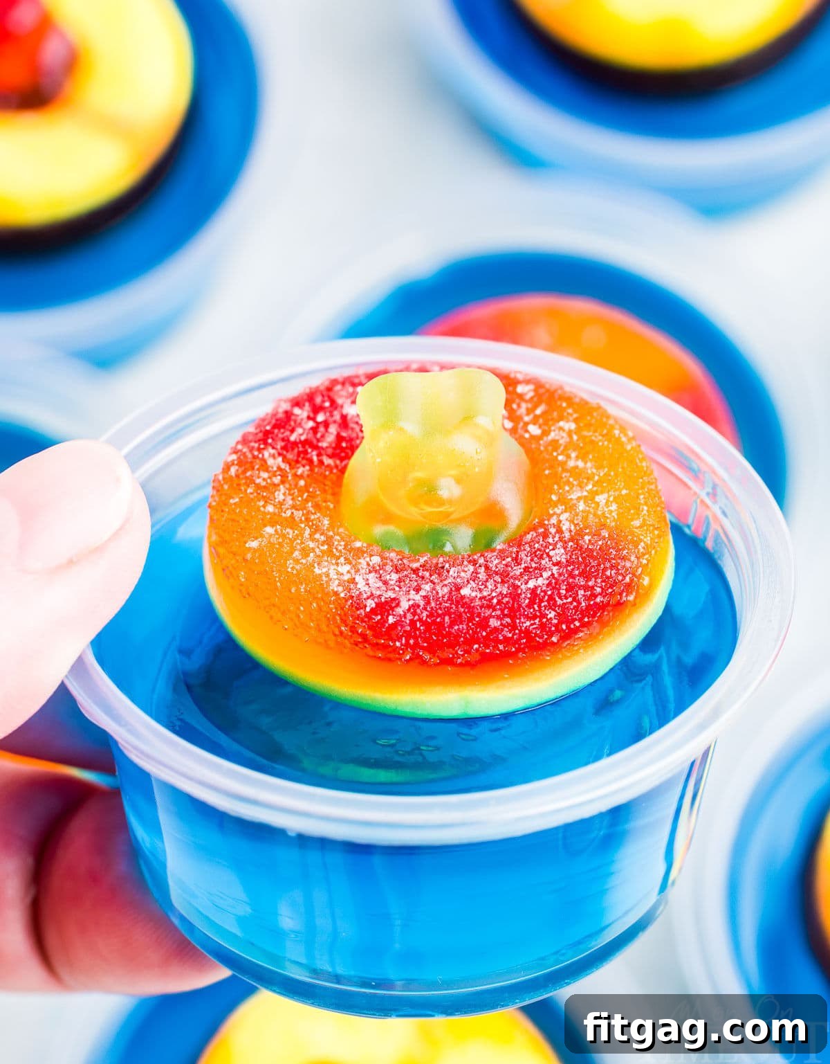Poolside Jell-O Jiggle 5 A hand holds a single vibrant blue jello shot, featuring a gummy bear perched on a peach ring floatie, against a backdrop of more festive jello shots.