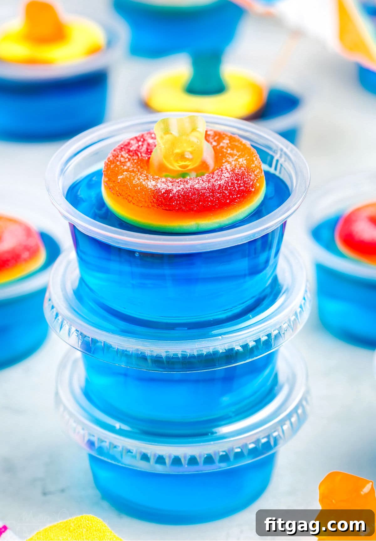 Poolside Jell-O Jiggle 6 Three blue jello shots stacked with clear plastic lids, the top shot beautifully adorned with a peach ring and a gummy bear, ready for a party.
