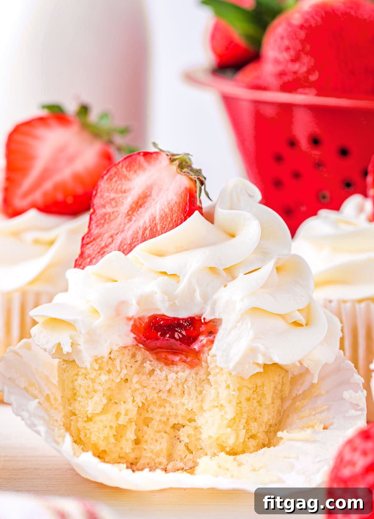 A vanilla cupcake with a visible strawberry jam filling and a swirl of buttercream frosting, topped with half a fresh strawberry. A bite has been taken, revealing the jam inside.