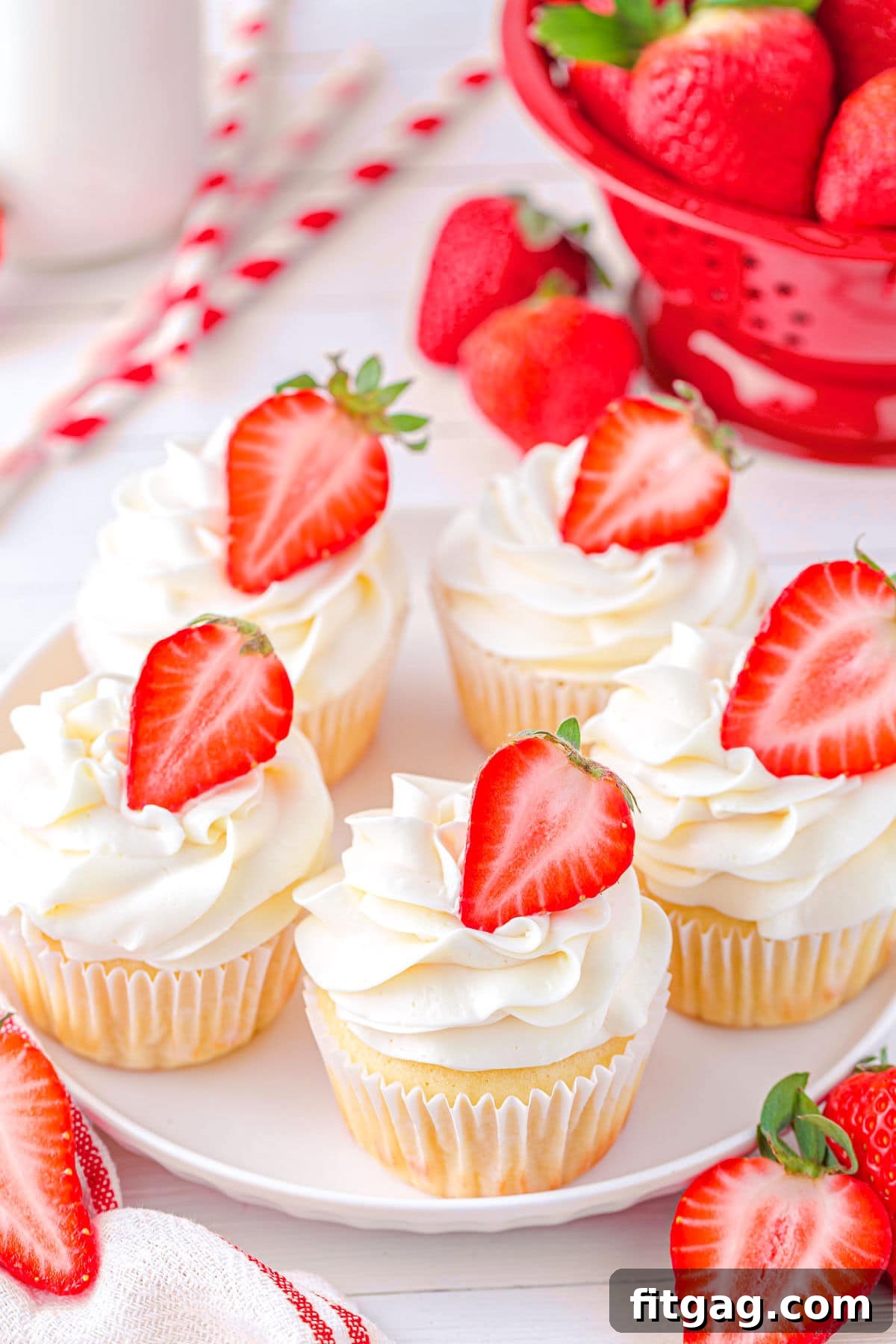 A top-down view of five vanilla cupcakes with strawberry jam filling and vanilla buttercream frosting, each garnished with half a fresh strawberry, arranged on a white plate.