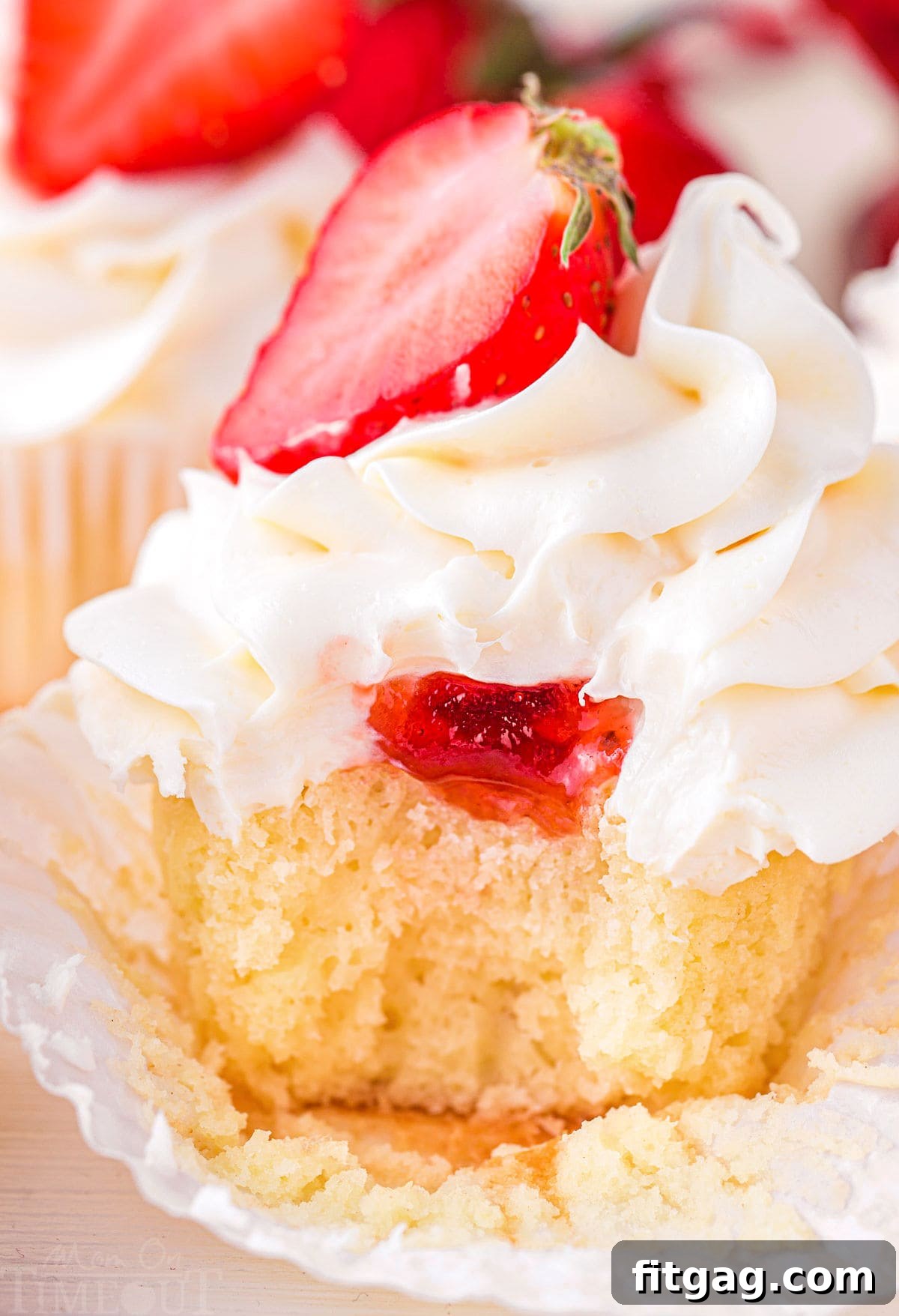 A close-up view of a strawberry shortcake cupcake, revealing the generous strawberry jam filling beneath a swirl of vanilla buttercream frosting.
