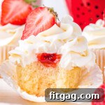 vanilla cupcake with strawberry filling and buttercream frosting. topped with half of a fresh strawberry. cupcake has a bite taken so you can see the strawberry jam on the inside.