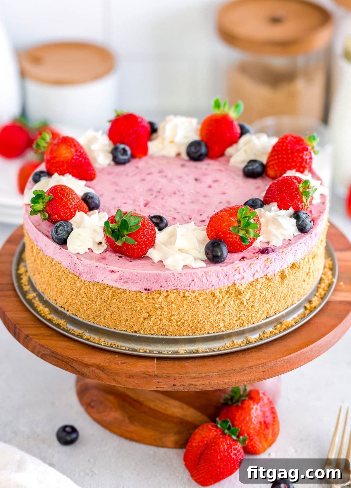 A vibrant No Bake Berry Cheesecake adorned with fresh blueberries and strawberries, topped with fluffy whipped cream, displayed on an elegant wooden cake stand surrounded by scattered berries.
