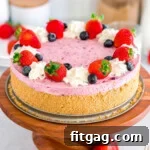 berry cheesecake topped with fresh blueberries and strawberries and whipped cream on a wood cake stand with more berries scattered around the base of the stand.