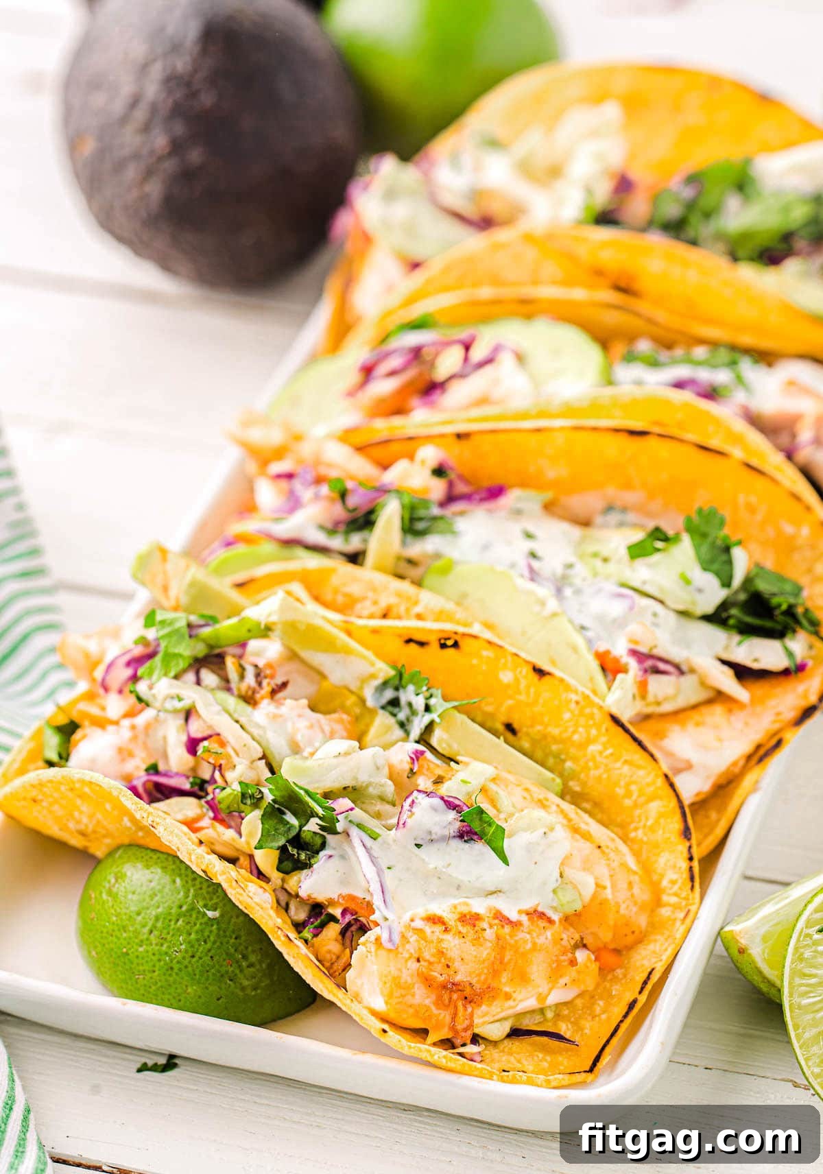 Three perfectly assembled fish tacos on a pristine white platter, generously topped with creamy fish taco sauce and fresh cilantro. Bright green lime wedges are elegantly placed beside the tacos, with a blurred avocado in the background suggesting even more fresh ingredients.