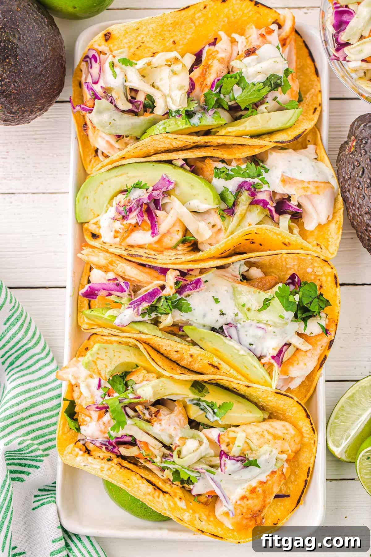 A top-down view of four artfully arranged fish tacos on a sleek white rectangular plate, garnished with fresh avocado slices, vibrant cilantro, and zesty lime wedges strategically placed around the presentation.