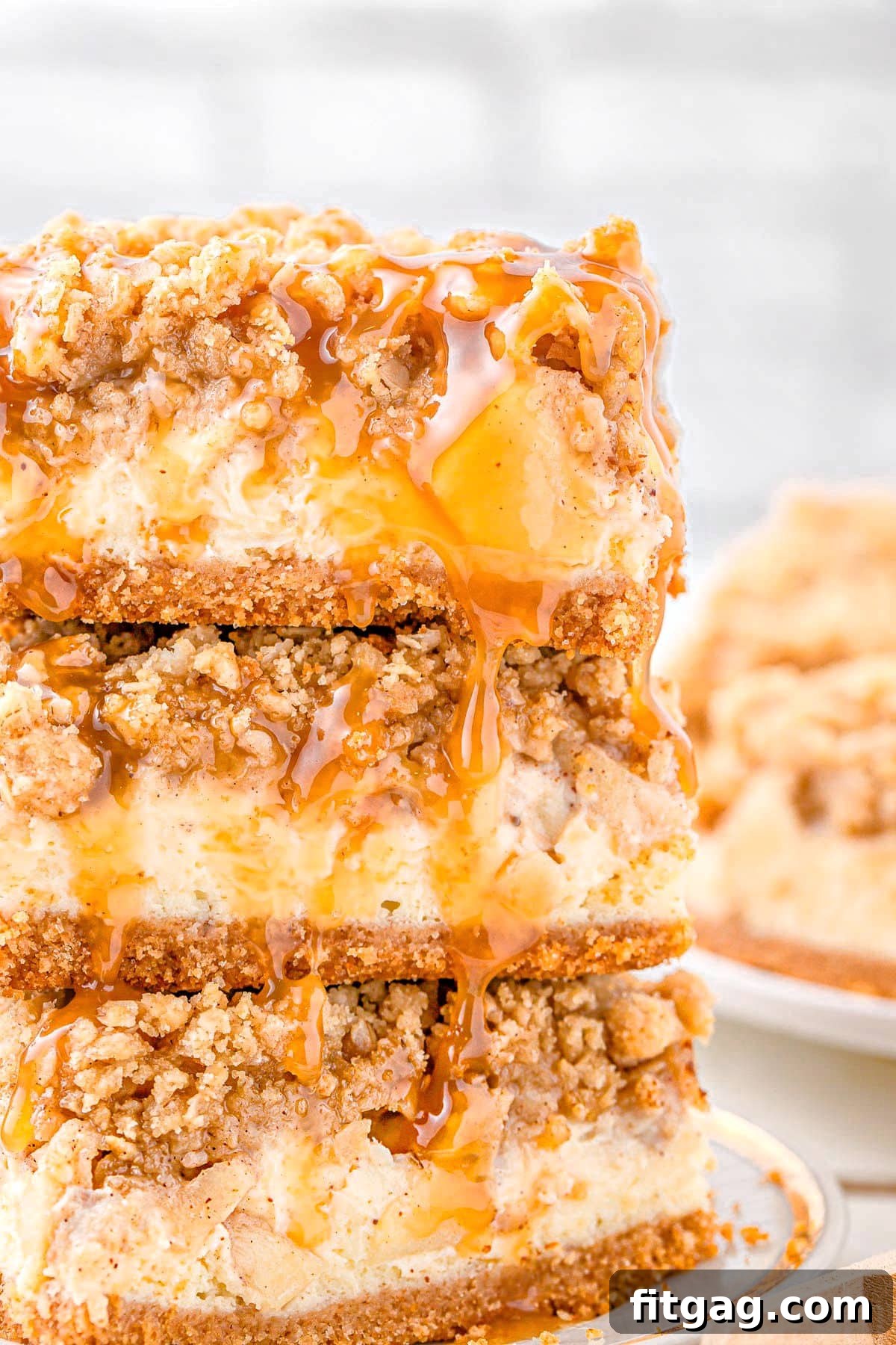 Three golden-brown caramel apple cheesecake bars stacked neatly on a white plate with a subtle gold rim. A generous drizzle of rich caramel sauce cascades over the top, highlighting the crumbly texture and inviting layers.